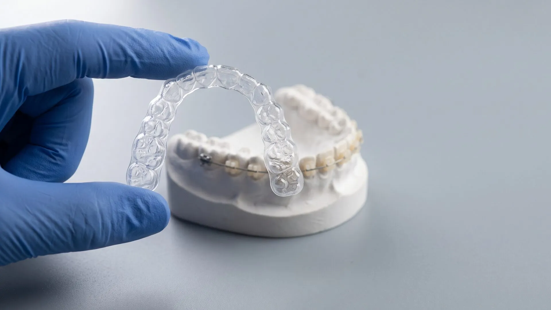A clear orthodontic aligner held in a gloved hand above a dental model fitted with ceramic braces