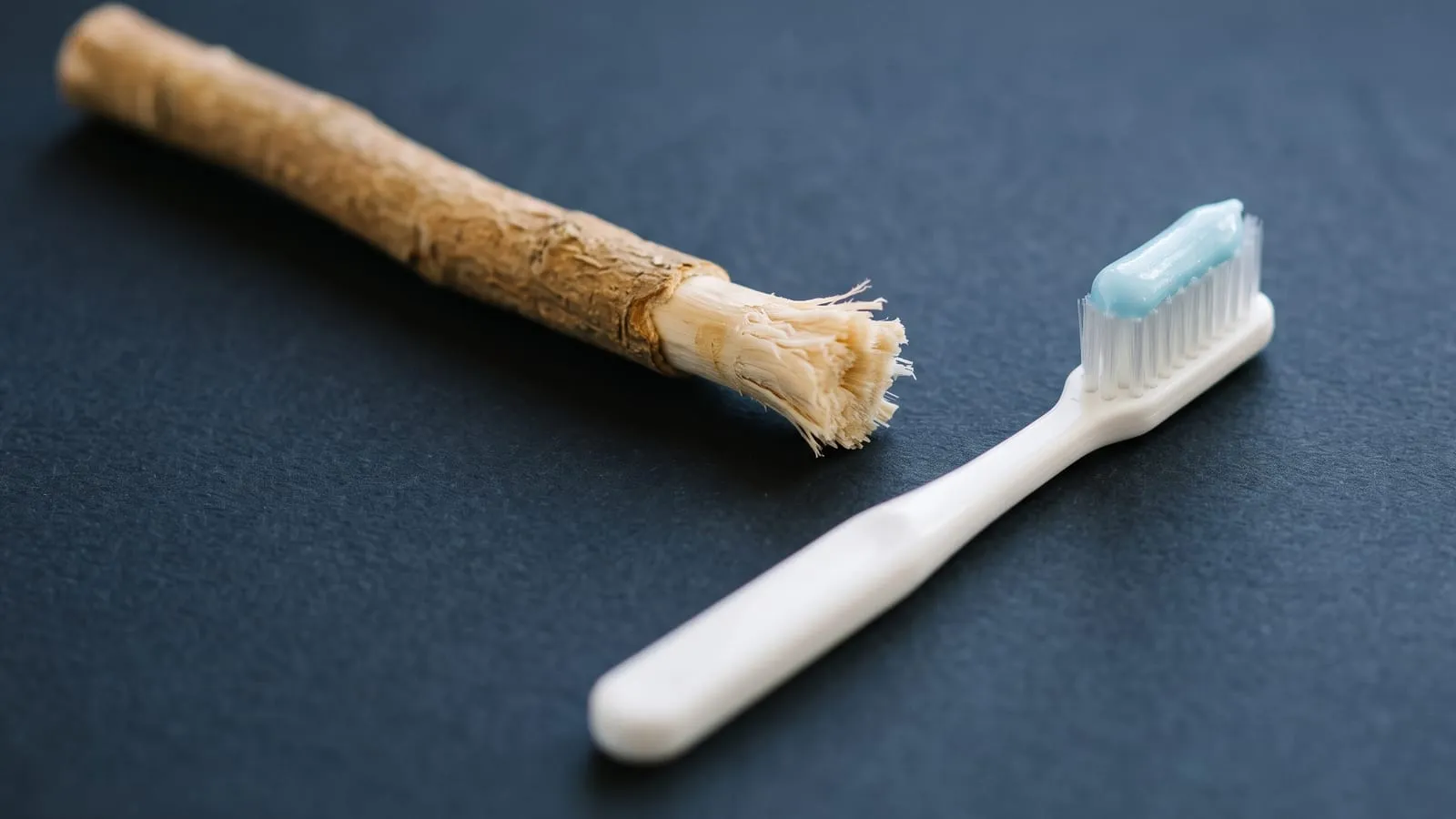 A miswak chewing stick with its bark peeled back and its tip chewed into pale frayed fibres, lying on a dark slate surface beside a white toothbrush with blue toothpaste on the bristles