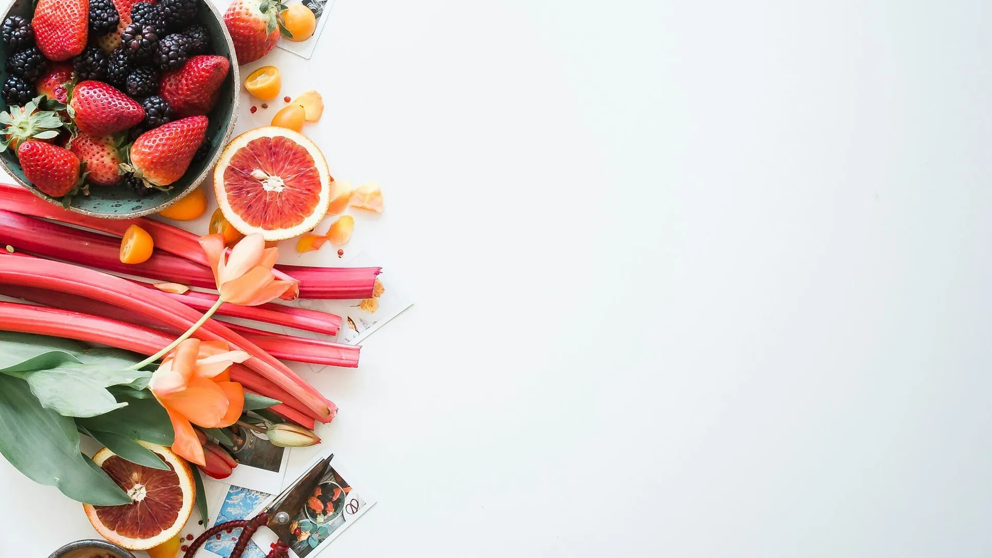 Fresh berries, citrus fruit, rhubarb and leafy greens laid out on a white surface